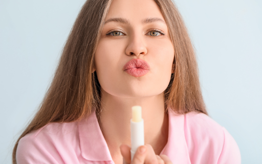 A young woman with long brown hair puckers her lips and holds up a tube of lip balm, representing daily lip care
