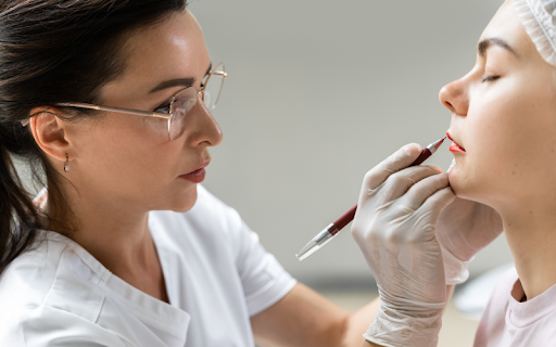 A cosmetic tattoo artist in glasses carefully outlines a client's lips with a pen